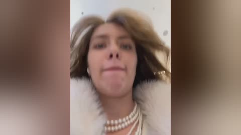 Media: Video of a woman with light skin, long blonde hair, and a white fur-trimmed coat, wearing a pearl necklace, with a blurred background.