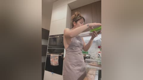 Media: Video of a woman in a modern kitchen, wearing a light apron, stirring a green liquid in a pot. Wooden cabinets, black oven, and a hanging towel are visible in the background.