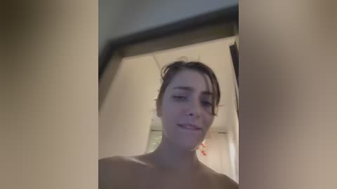 Media: Video of a young woman with light skin and dark hair, wearing a black top, looking at herself in a mirror. Background shows a bathroom with white tiles and a red towel.