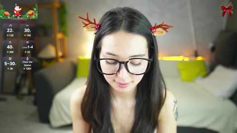 Video of a slender Asian woman with long black hair, wearing red antlers and glasses, smiling softly. Background shows a modern bedroom with a bed, green pillows, and a yellow lamp.