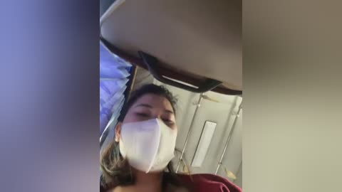 Media: A video of a woman with light skin, wearing a white face mask, looking downward while seated on a train, with blurred background showing metal bars and a window.