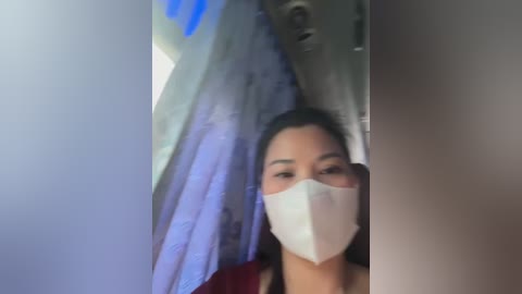 Media: Video of a woman with light brown skin and straight black hair, wearing a white surgical mask, standing indoors in a dimly lit room with white curtains and a blue light source.