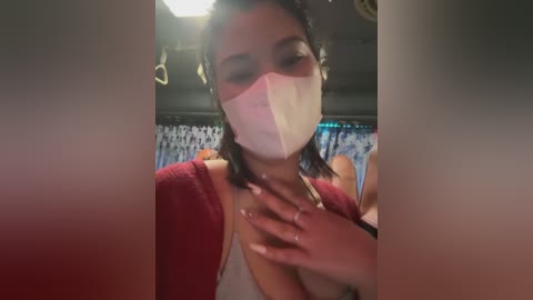 Media: Video of a woman in a red cardigan, wearing a white mask, with a blurred background featuring blue curtains.