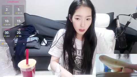 Media: Video of an East Asian woman with long black hair, wearing a white t-shirt, sitting at a table with a straw cup and banana, in a modern, minimalistic room with a black couch.