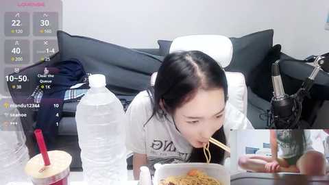 Media: Video of an Asian woman with long black hair, eating noodles from a bowl on a table, with a glass of water and a red straw cup. Background includes a black couch, clothes, and a microphone.