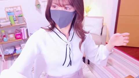 Media: Video of an East Asian woman with long brown hair, wearing a white hoodie, black mask, and gray gloves, standing in a cozy, well-lit room with shelves of cleaning products, a white couch, and a wooden door.