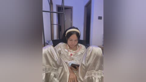 Media: Video of a young Asian woman with dark hair, wearing a white satin robe, sitting on a black chair, reading a book, in a dimly lit hallway.