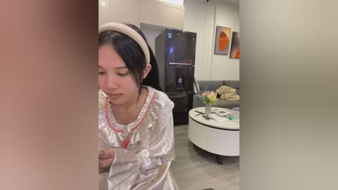 Media: Video of a young Asian woman with long black hair, wearing a white dress, looking down, standing in a modern living room with a gray sofa, white coffee table, and abstract art on the wall.