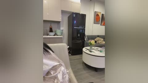 Media: Video of a modern, minimalist living room with a black fridge, white sofa, and grey floor. Artwork and plants adorn the walls, adding a touch of color.