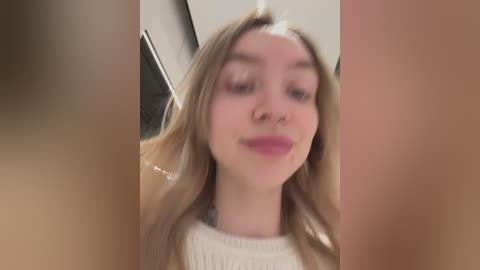 Media: Video of a young, light-skinned woman with long, blonde hair and fair skin, wearing a white sweater, taken from a low angle, with blurred, dark-haired individuals in the background.