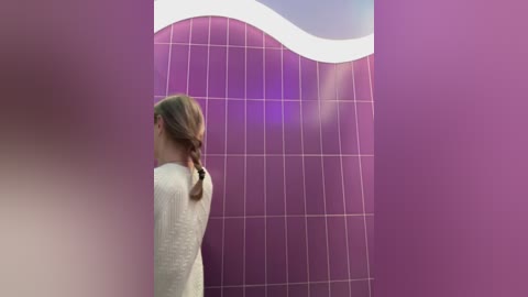 A video of a young woman with light skin and blonde hair in a braid, wearing a white sweater, standing in front of a large, purple tiled wall with a curved, white wave design. The background is soft-focus, emphasizing the wall's texture and color.