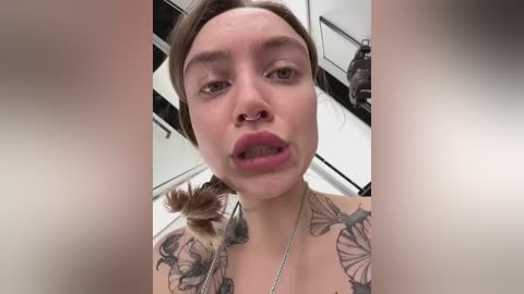 A close-up video of a young woman with pale skin, brown hair, and numerous tattoos including butterflies on her shoulders. She wears a nose ring and has a slight pout, taken indoors.