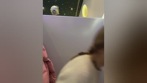 Media: Video of a child with long brown hair wearing a pink jacket, looking through a window at a moon and planet model in a space-themed room.