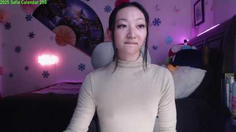 Media: A video of a woman with light skin, straight black hair, and wearing a beige turtleneck sweater, standing in a purple-lit bedroom with a pink flamingo lamp, a plush toy, and a bed with a white pillow.