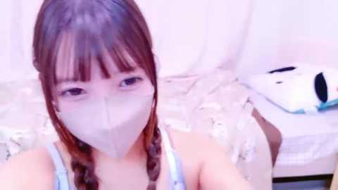 Media: A young Asian woman with braided brown hair and bangs, wearing a light blue tank top, white mask, and a floral bedsheet in a softly lit bedroom.