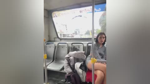 Media: Video of a woman in a gray jacket and red shorts sitting on a bus seat, surrounded by other passengers, with a clear window view of a cityscape.