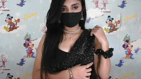 Media: Video of a young woman with long black hair, wearing a black mask, black blouse, pearl necklace, and black scrunchie, against a Disney-themed backdrop with colorful characters.