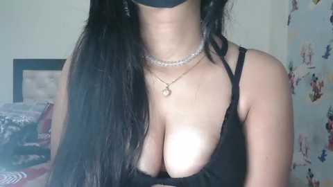 Media: Video of a woman with long black hair, wearing a black bra, necklace, and black face mask, in a dimly lit bedroom with floral wallpaper and a bed in the background.