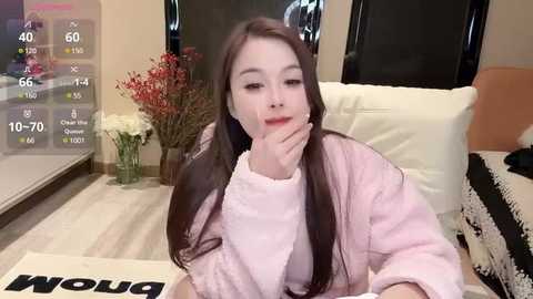 Media: Video of an East Asian woman with long dark hair, wearing a pink robe, sitting on a white couch in a modern living room with a TV, flowers, and a digital clock displaying 40\u00b0C.