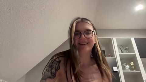 Media: Video of a smiling, glasses-wearing woman with long blonde hair and a tattoo sleeve, standing indoors near white shelves and beige walls.