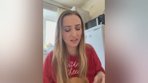 Media: Video of a young, light-skinned woman with long, straight blonde hair wearing a red t-shirt, sitting in a kitchen, looking at a stuffed teddy bear, with a microwave and fridge in the background.