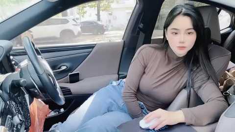 Media: Video of a young East Asian woman with long black hair, wearing a tight brown turtleneck and light blue jeans, seated in a car, holding a mouse, with a blurred outdoor background.