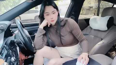 Media: A video of an Asian woman with long black hair, fair skin, wearing a tight, brown, long-sleeved top and white shorts, sitting in the passenger seat of a car, looking tired and thoughtful.