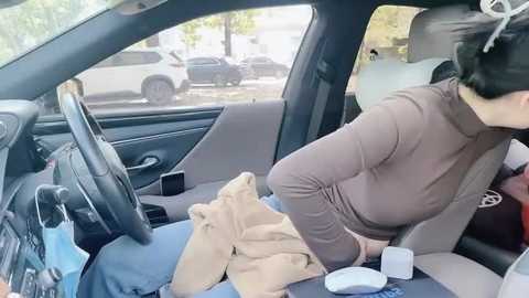 Media: Video of a woman with dark hair and light skin in a car, wearing a brown turtleneck sweater and beige coat, leaning over a child's car seat, with a white cup and smartphone on the seat.