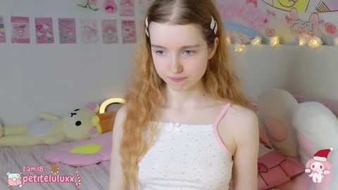 Media: Video of a young, pale-skinned, blonde-haired girl with long, wavy hair in a white, polka-dotted tank top, sitting on a bed with plush toys and fairy lights in the background.