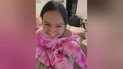 Media: Video of a young Asian woman with long black hair, wearing a pink tie-dye hoodie, smiling broadly on a bed in a cluttered room with a TV and various items in the background.