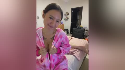 Media: A video of an Asian woman with dark hair, wearing a pink tie-dye robe, standing in a messy bedroom.