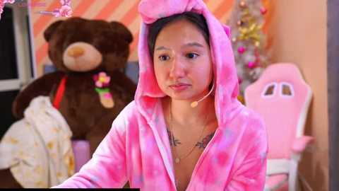 Media: Video of an Asian woman with dark hair in a pink, tie-dye teddy bear onesie, wearing a necklace, standing in a cozy room with a brown teddy bear and a Christmas tree.