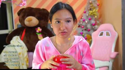 Media: Video of an Asian girl with straight black hair, wearing a pink tie-dye hoodie, holding a red apple. Background includes a teddy bear, a pink gaming chair, and a decorated Christmas tree. Text overlays include \"my/our cute little\" and \"my/our cute little...\