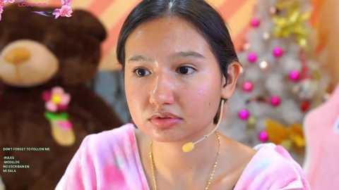 Media: Video of a young Asian woman with straight black hair, wearing a pink tie-dye shirt and gold necklace, sitting in a room with a pink and white Christmas tree and stuffed toys.