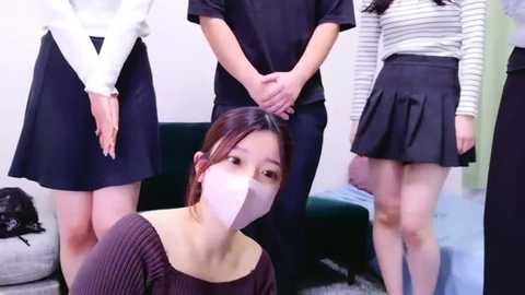 Media: Video of four Asian women in modern, casual outfits, two in black dresses, one in a striped top, and one in a sweater, standing in a room with a green couch and a grey carpet.