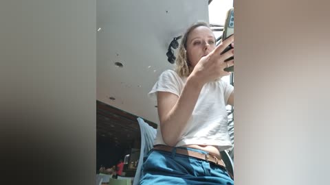 Media: Video of a young woman with blonde hair, wearing a white t-shirt and blue jeans, taking a selfie in a mirror. The background shows a cluttered room with a wooden desk and a blue chair.