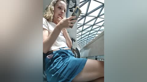 Media: Video of a blonde woman with light skin and shoulder-length hair, wearing a white t-shirt and blue denim skirt, sitting on a chair in a modern, sunlit room with a glass wall and grid-patterned skylight.