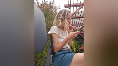 Media: Video of a young blonde woman in a white t-shirt and blue skirt, eating an ice cream cone, in a garden with a red metal trellis and green plants in the background.