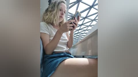 Media: Video of a blonde woman in a white t-shirt and denim shorts, reading a book, seated in a dimly lit, angular room with a large, geometric skylight window.