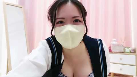 Media: Video of an East Asian woman in a white face mask, black vest over a low-cut blouse, and blue-and-white sailor collar, in a pink room with a white dresser.