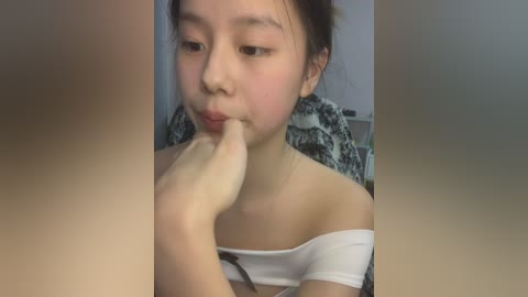 Media: Video of a young Asian woman with light skin, brown hair, and a white off-shoulder top, biting her finger in a contemplative manner. Background shows a floral-patterned chair.