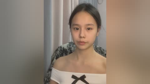 Media: Video of an Asian woman with straight, dark hair, wearing a white off-shoulder top with a black bow, standing indoors.