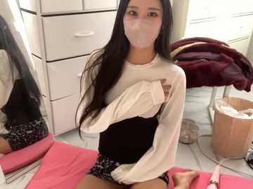 Media: Video of a young Asian woman with long black hair, wearing a white long-sleeved top and black shorts, sitting on a pink mat in a messy room with white drawers, a cat tower, and a basket.