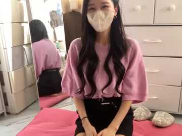 Media: Video of an East Asian woman with long black hair, wearing a light pink t-shirt and black skirt, seated on a pink mat in a room with white furniture and a mirror.