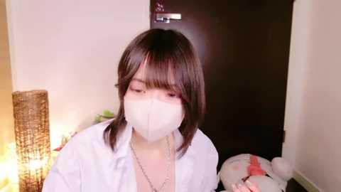 Media: Video of an Asian woman with shoulder-length dark hair, wearing a white surgical mask, sitting in a dimly-lit room with a wicker chair and a teddy bear.