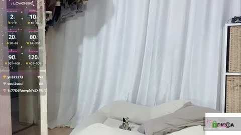 Media: Video of a bedroom with white curtains partially drawn, showing a bed with white sheets. A live video overlay displays time, temperature, and weather information.