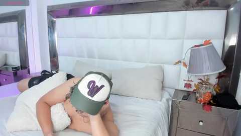 Media: A video of a woman in a white lace bra, wearing a green and white baseball cap, lying on a white bed with a tufted headboard, in a modern bedroom with a mirrored wall, a nightstand with a lamp and flowers, and purple lighting.