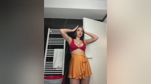 Media: Video of a woman with long dark hair, wearing a red crop top and yellow skirt, standing in a modern bathroom with white door and black tiled walls, holding a white towel.