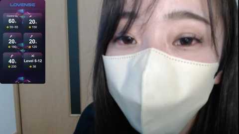 Media: Video of an East Asian woman with straight black hair and fair skin, wearing a white face mask, looking directly at the camera. Background includes a Lovense touchscreen device displaying various percentages.