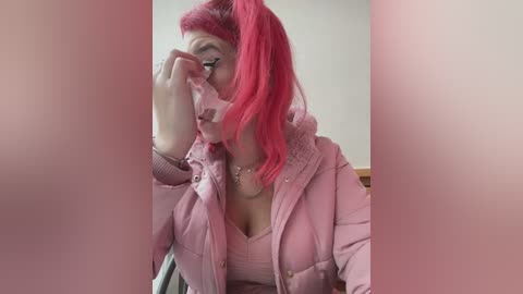 Media: Video of a young woman with bright red hair, wearing a pink quilted jacket, adjusting her glasses. She has a fair complexion and is indoors, possibly in a living room.